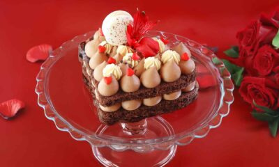 Heart‑shaped chocolate cake topped with milk chocolate mousse, cream swirls and red accents, displayed on a glass stand against a red background by AL HALLAB.