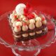 Heart‑shaped chocolate cake topped with milk chocolate mousse, cream swirls and red accents, displayed on a glass stand against a red background by AL HALLAB.