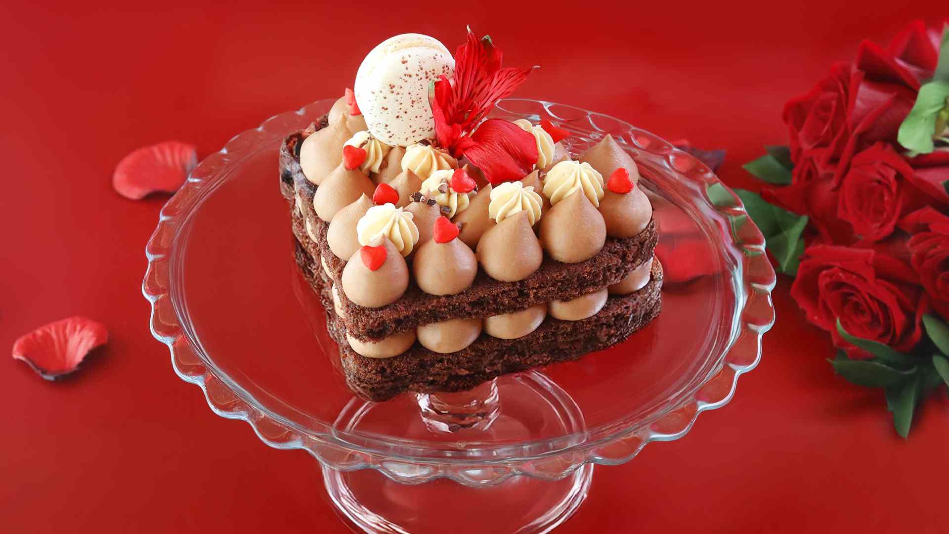 Heart‑shaped chocolate cake topped with milk chocolate mousse, cream swirls and red accents, displayed on a glass stand against a red background by AL HALLAB.