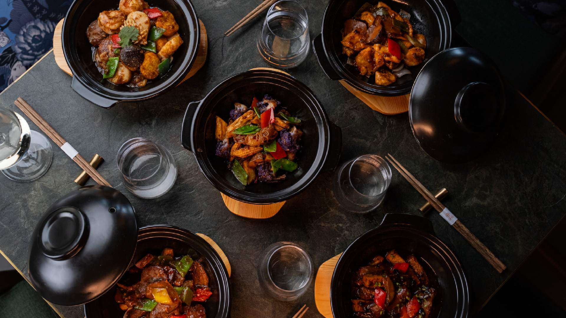 Five traditional Chinese claypot dishes served on a dark stone table at SHI restaurant, featuring vibrant vegetables, tender meats, and rich sauces in black claypots with wooden bases, surrounded by chopsticks, empty glasses, and lids