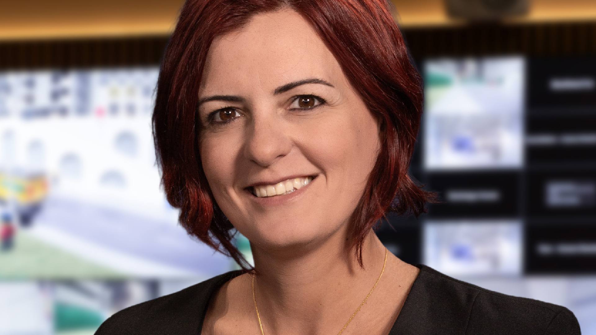 Close-up view of a person wearing a black outfit with a gold necklace, standing in front of a blurred background showing multiple video surveillance screens in a control room environment