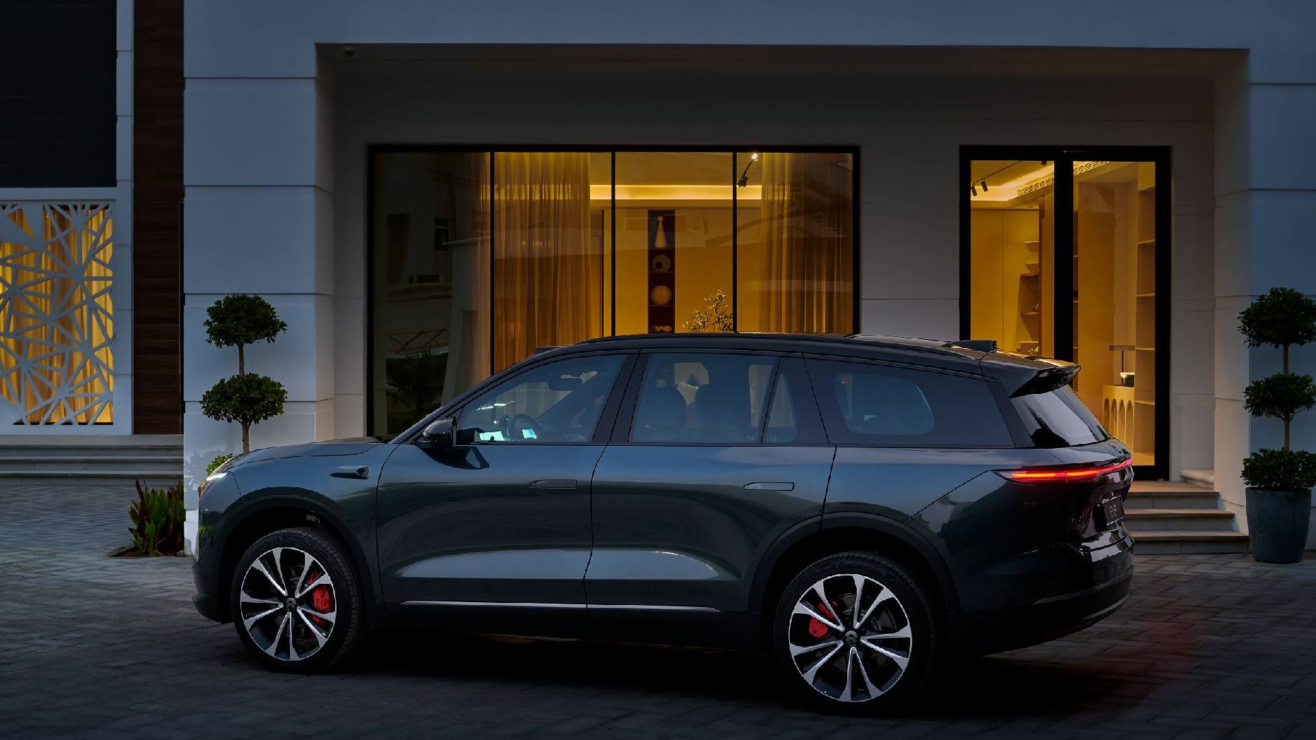Side view of a dark gray NIO electric SUV parked on a paved driveway in front of a modern house with large glass doors and warm interior lighting.