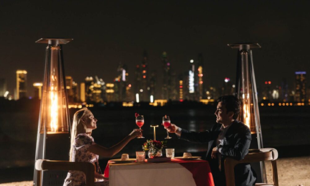 Romantic seaside dinner setup at Villamoré with two guests toasting glasses of red wine under the night sky, flanked by outdoor heaters, with Dubai city skyline illuminated in the background.