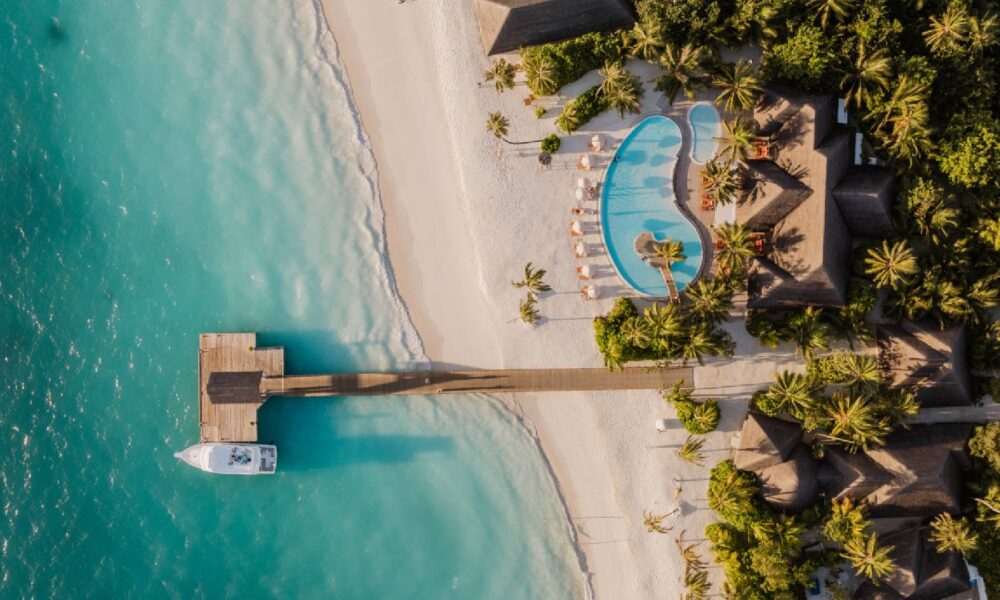 Aerial view of Sun Siyam Iru Veli resort showing turquoise ocean, white sandy beach, wooden pier with a docked boat, and a lagoon-shaped swimming pool surrounded by sun loungers and tropical palm trees. Would you like me to also provide an SEO-friendly caption and optimized image file name for WordPress upload?