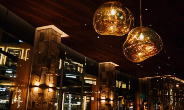 Warm interior view of 99 Sushi Bar & Restaurant featuring two modern amber glass pendant lights hanging from a wooden ceiling, with stone columns and softly lit wall sconces creating an elegant, upscale dining ambiance.