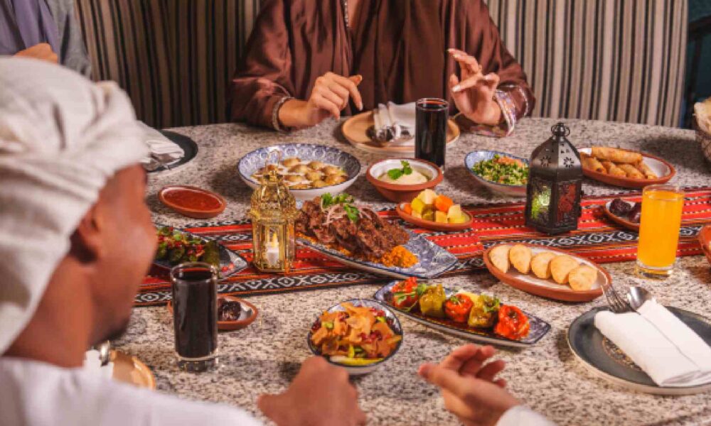 Guests in traditional Middle Eastern attire share a Ramadan iftar table at BESH TURKISH KITCHEN, with assorted Turkish mezze, grilled kebabs, bread, juices, and lanterns on a patterned table.