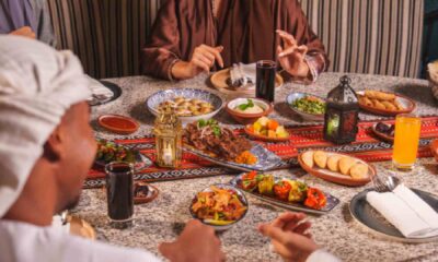 Guests in traditional Middle Eastern attire share a Ramadan iftar table at BESH TURKISH KITCHEN, with assorted Turkish mezze, grilled kebabs, bread, juices, and lanterns on a patterned table.