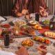 Guests in traditional Middle Eastern attire share a Ramadan iftar table at BESH TURKISH KITCHEN, with assorted Turkish mezze, grilled kebabs, bread, juices, and lanterns on a patterned table.
