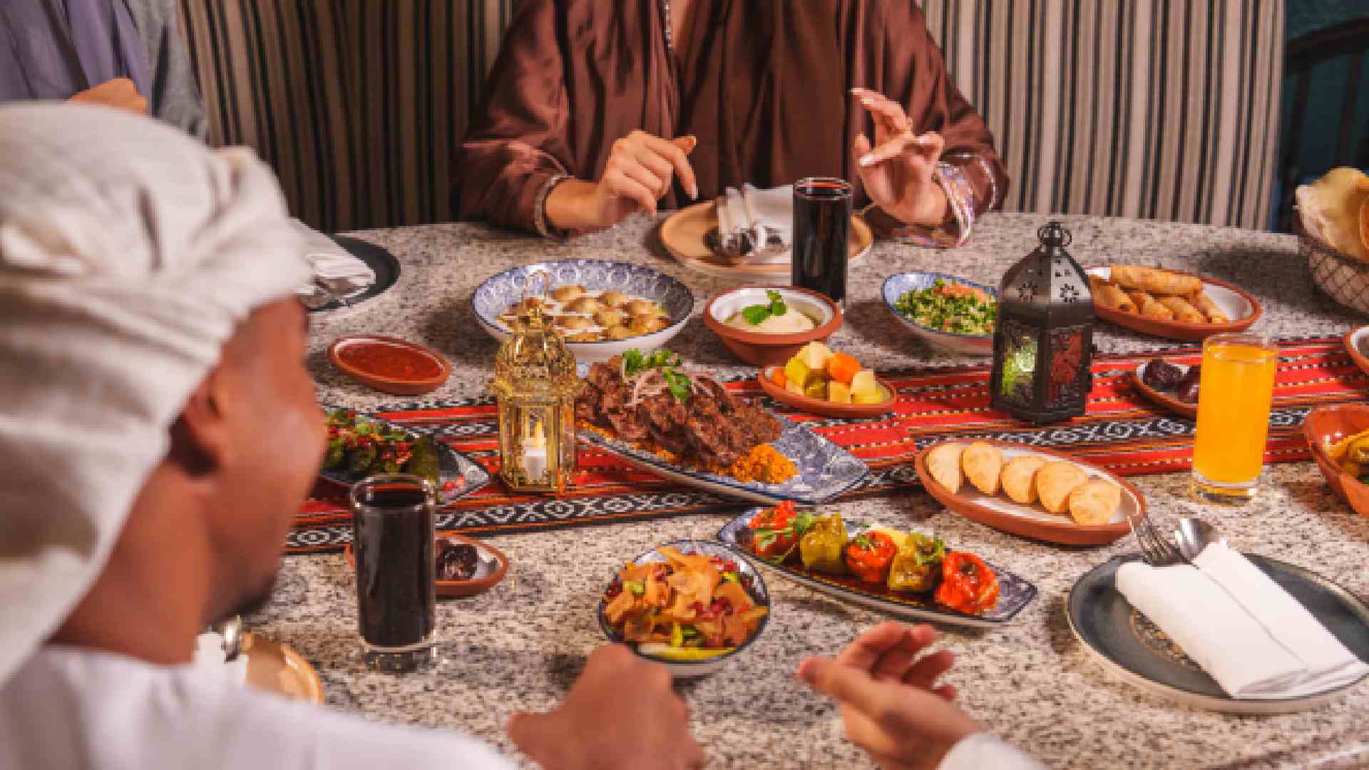Guests in traditional Middle Eastern attire share a Ramadan iftar table at BESH TURKISH KITCHEN, with assorted Turkish mezze, grilled kebabs, bread, juices, and lanterns on a patterned table.