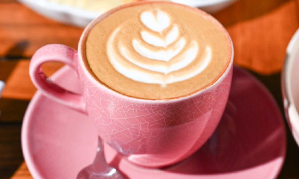A freshly prepared latte topped with delicate leaf‑style latte art is served in a pink cup and saucer at COUNTER CULTURE CAFÉ. Placed on a wooden table, the coffee is accompanied by a breakfast platter in the background, featuring a selection of light bites, creating a relaxed café setting ideal for coffee breaks or casual dining.