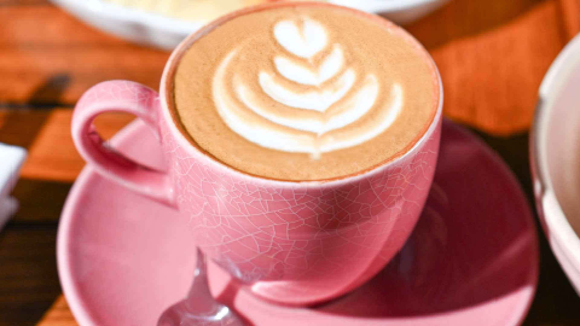 A freshly prepared latte topped with delicate leaf‑style latte art is served in a pink cup and saucer at COUNTER CULTURE CAFÉ. Placed on a wooden table, the coffee is accompanied by a breakfast platter in the background, featuring a selection of light bites, creating a relaxed café setting ideal for coffee breaks or casual dining.