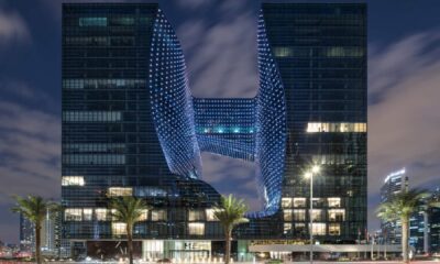 Night view of MELIÁ DUBAI’s Opus building showing two illuminated towers connected by a central sky bridge, with palm trees in the foreground and Dubai skyline in the background.