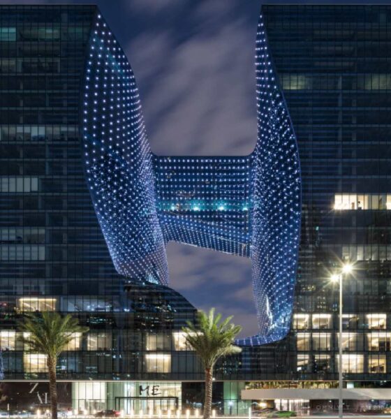 Night view of MELIÁ DUBAI’s Opus building showing two illuminated towers connected by a central sky bridge, with palm trees in the foreground and Dubai skyline in the background.