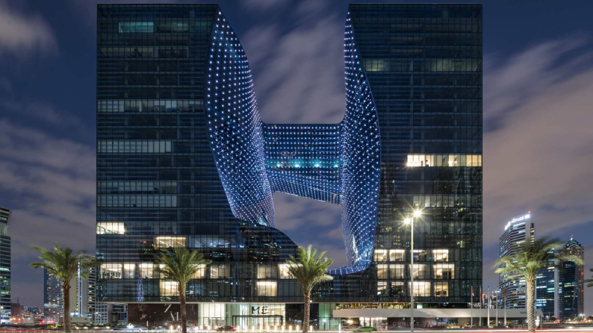 Night view of MELIÁ DUBAI’s Opus building showing two illuminated towers connected by a central sky bridge, with palm trees in the foreground and Dubai skyline in the background.