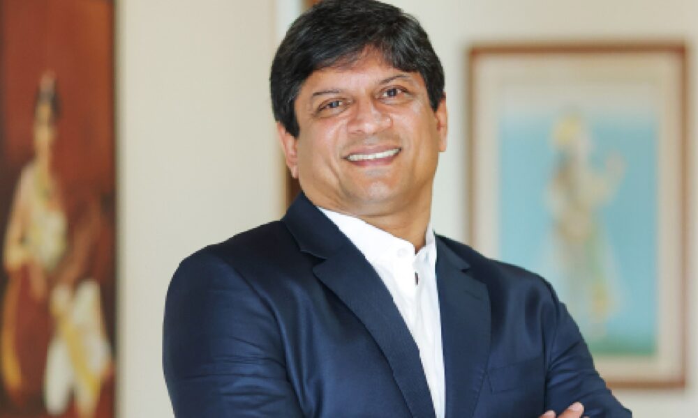 Person wearing a dark blue formal suit with a white shirt, standing indoors with arms crossed. The background features two framed paintings on a light-colored wall.