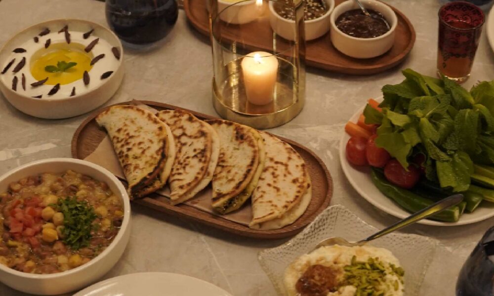 Overhead view of a BAB EL BAHR Suhoor table featuring freshly baked Arabic bread, scrambled eggs, hummus, foul medammas, labneh, pita bread, olives, herbs, and mixed Levantine dishes served with tableware and a lit candle.