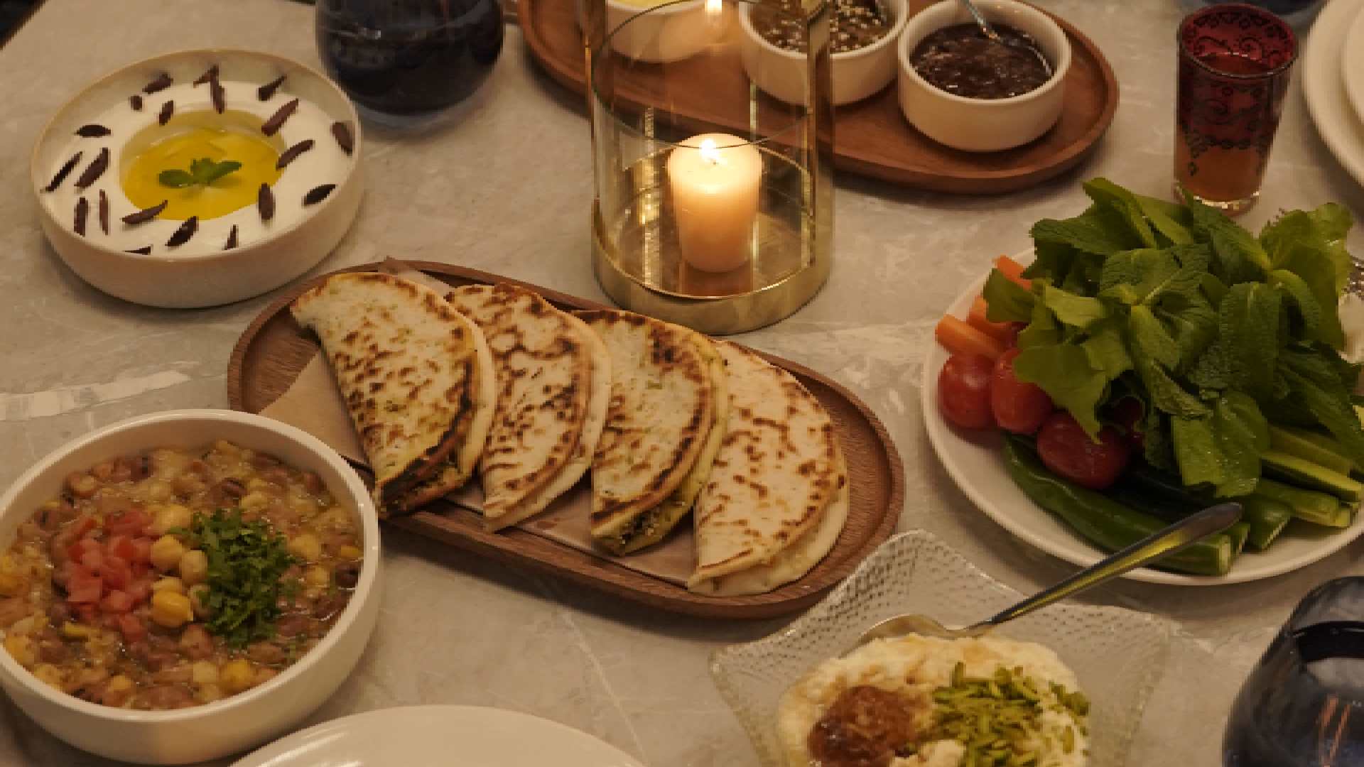 Overhead view of a BAB EL BAHR Suhoor table featuring freshly baked Arabic bread, scrambled eggs, hummus, foul medammas, labneh, pita bread, olives, herbs, and mixed Levantine dishes served with tableware and a lit candle.