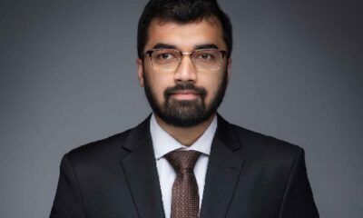 A person wearing a dark formal suit and tie stands against a plain grey studio background, holding their hands together in a composed, professional pose.