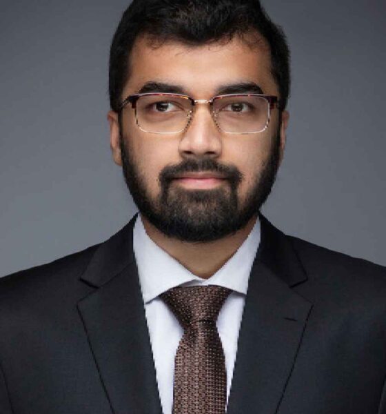 A person wearing a dark formal suit and tie stands against a plain grey studio background, holding their hands together in a composed, professional pose.