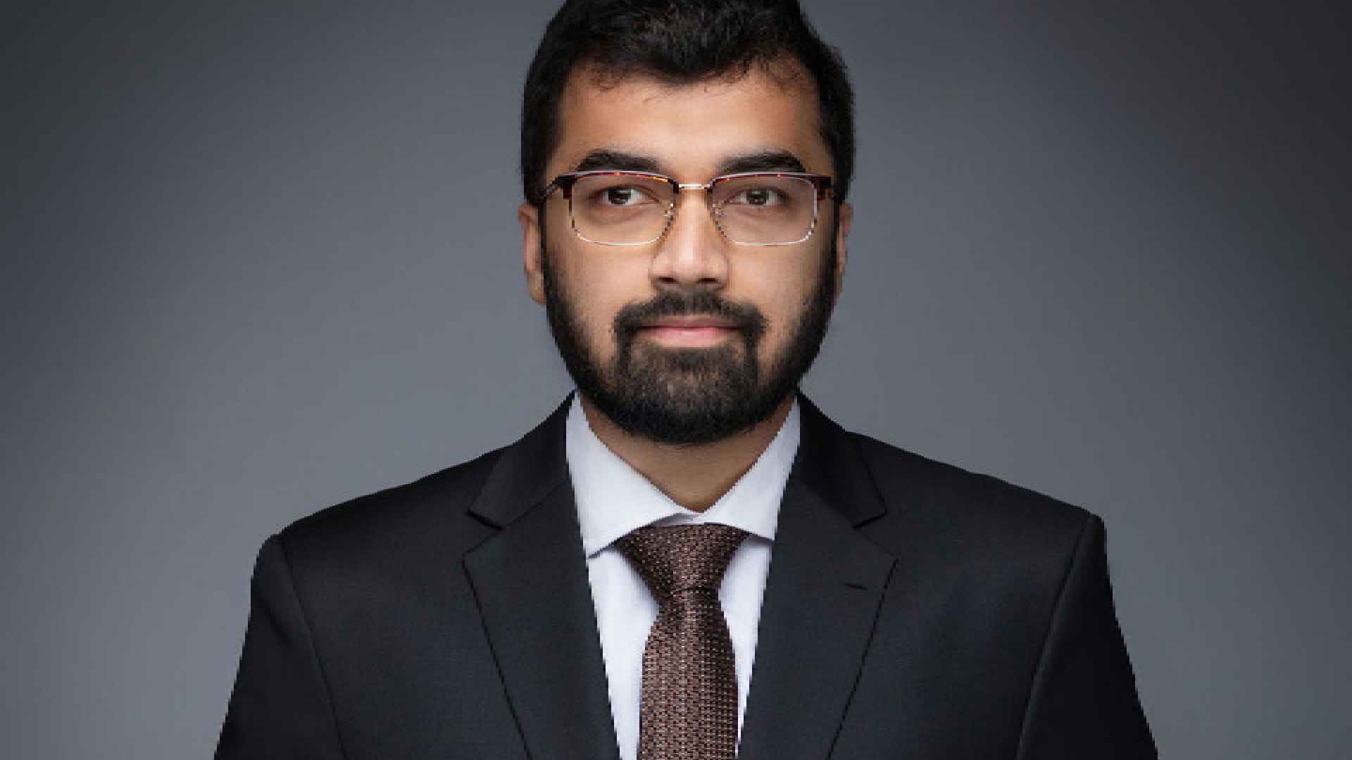 A person wearing a dark formal suit and tie stands against a plain grey studio background, holding their hands together in a composed, professional pose.
