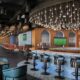 A modern pub interior featuring a curved bar with high stools, a large display of spirits, and warm ambient lighting with hanging pendant bulbs. The space includes teal and brown seating, long rows of tables, large windows along one side, and wall‑mounted screens displaying a sports match.