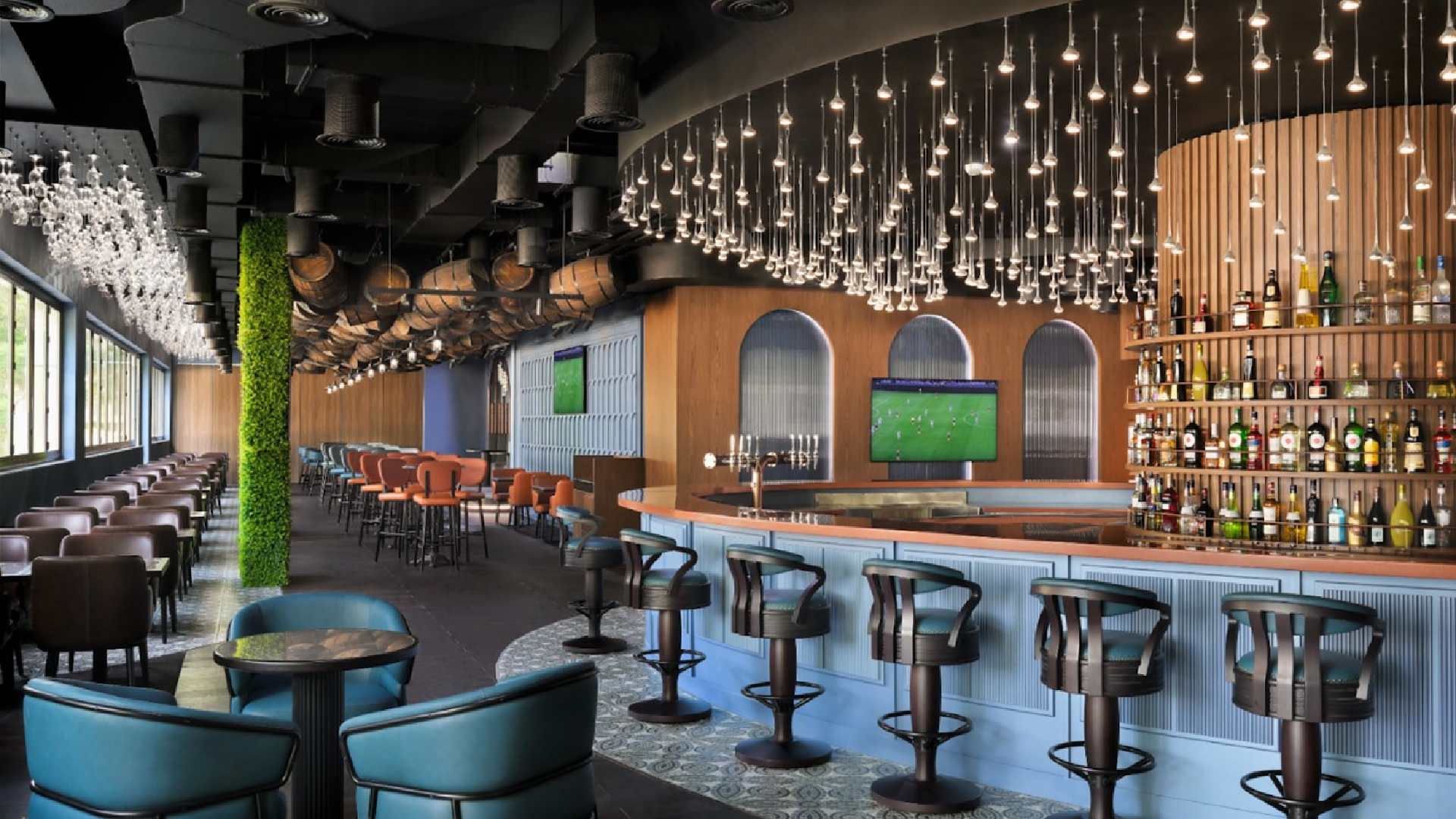 A modern pub interior featuring a curved bar with high stools, a large display of spirits, and warm ambient lighting with hanging pendant bulbs. The space includes teal and brown seating, long rows of tables, large windows along one side, and wall‑mounted screens displaying a sports match.