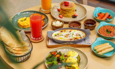 A table at Grazer Downtown set with a colorful breakfast spread, including pancakes topped with berries, eggs Benedict, scrambled eggs, baked beans, labneh, dips, grilled vegetables, bread, vegetable sticks, and two glasses of fresh juice, photographed through green plants in a bright indoor setting.