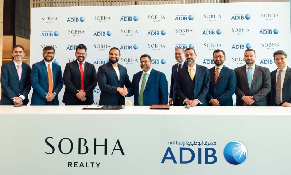 A group of executives from Sobha Realty and Abu Dhabi Islamic Bank stand together behind a signing table as representatives from both organizations shake hands in front of a branded backdrop displaying the Sobha Realty and ADIB logos, marking their partnership agreement.