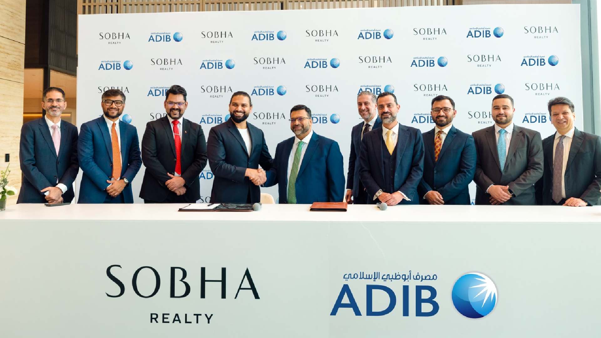 A group of executives from Sobha Realty and Abu Dhabi Islamic Bank stand together behind a signing table as representatives from both organizations shake hands in front of a branded backdrop displaying the Sobha Realty and ADIB logos, marking their partnership agreement.