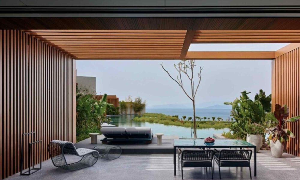 Covered outdoor terrace at Maxx Royal Bodrum Resort featuring a modern dining table and chairs, sun loungers facing a private infinity pool, wooden slatted pergola, lush landscaping, and panoramic sea view beyond the pool.