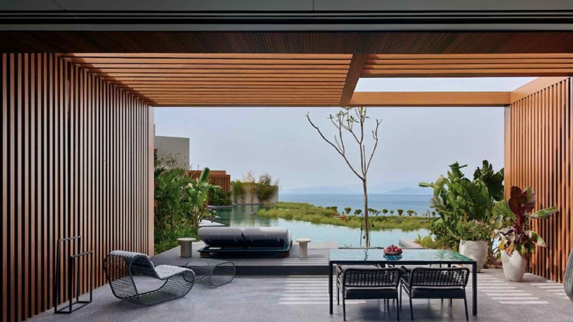 Covered outdoor terrace at Maxx Royal Bodrum Resort featuring a modern dining table and chairs, sun loungers facing a private infinity pool, wooden slatted pergola, lush landscaping, and panoramic sea view beyond the pool.