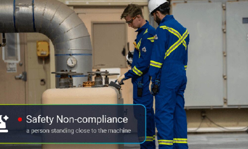 wo workers in blue safety uniforms with reflective stripes standing near industrial machinery and large piping, with an on-screen alert indicating “Safety Non-compliance: a person standing close to the machine