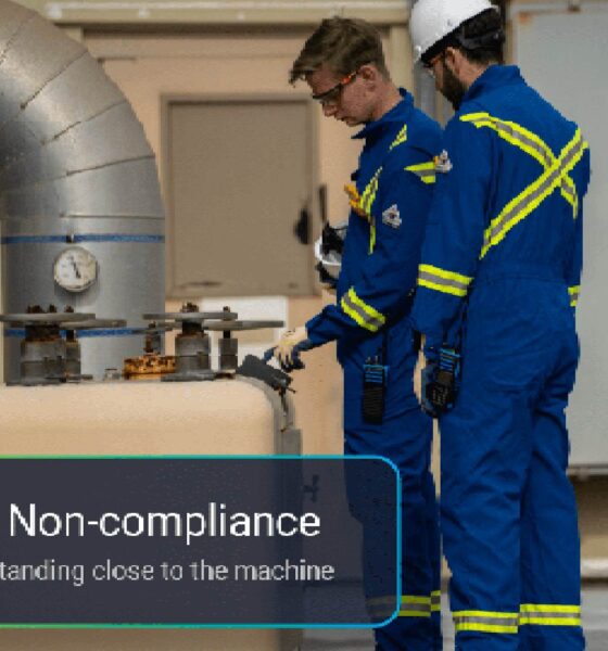 wo workers in blue safety uniforms with reflective stripes standing near industrial machinery and large piping, with an on-screen alert indicating “Safety Non-compliance: a person standing close to the machine