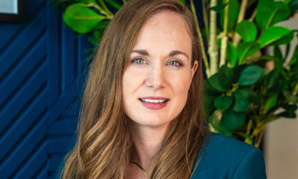 SharpMinds team member seated in a contemporary office setting, wearing a teal blazer over a white top, with textured wall panels and indoor plants in the background.