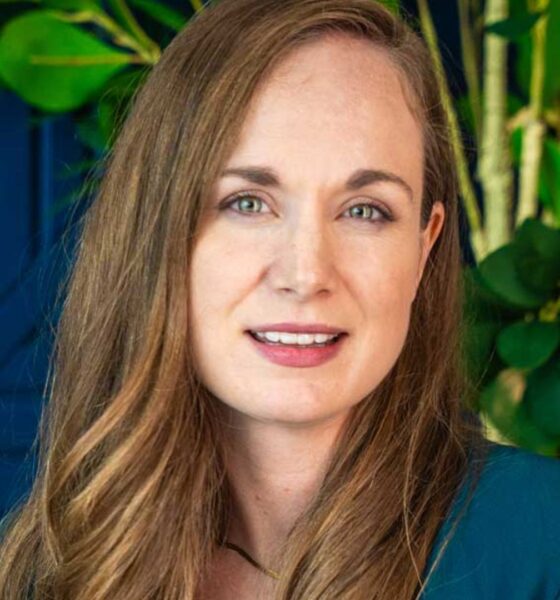 SharpMinds team member seated in a contemporary office setting, wearing a teal blazer over a white top, with textured wall panels and indoor plants in the background.