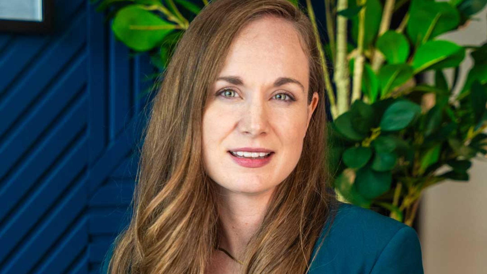 SharpMinds team member seated in a contemporary office setting, wearing a teal blazer over a white top, with textured wall panels and indoor plants in the background.