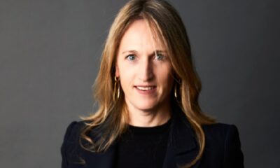 Professional studio portrait of a woman standing against a dark grey background, wearing a navy blazer over a black top and white trousers, with hands in pockets, photographed for Kerten Hospitality.