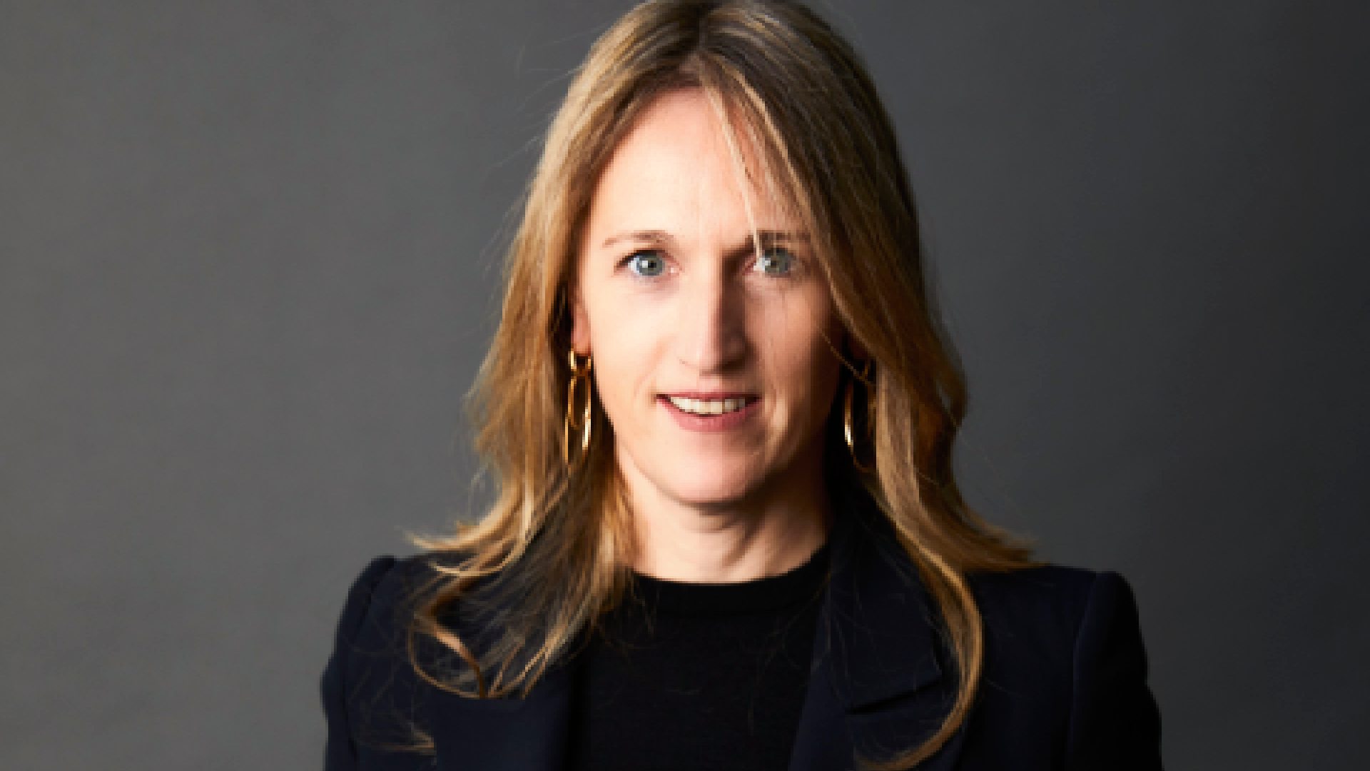 Professional studio portrait of a woman standing against a dark grey background, wearing a navy blazer over a black top and white trousers, with hands in pockets, photographed for Kerten Hospitality.