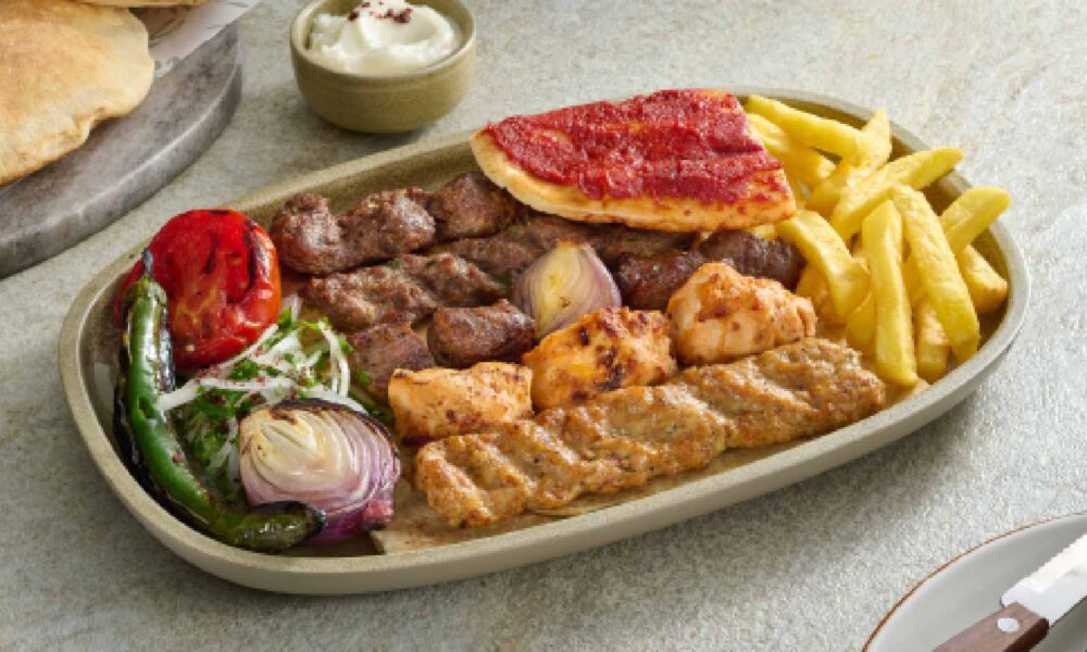 AL HALLAB mixed grill platter featuring grilled lamb cubes, chicken skewers, kafta, fries, grilled tomato, onions, flatbread, and garlic sauce on a serving plate.