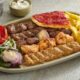 AL HALLAB mixed grill platter featuring grilled lamb cubes, chicken skewers, kafta, fries, grilled tomato, onions, flatbread, and garlic sauce on a serving plate.