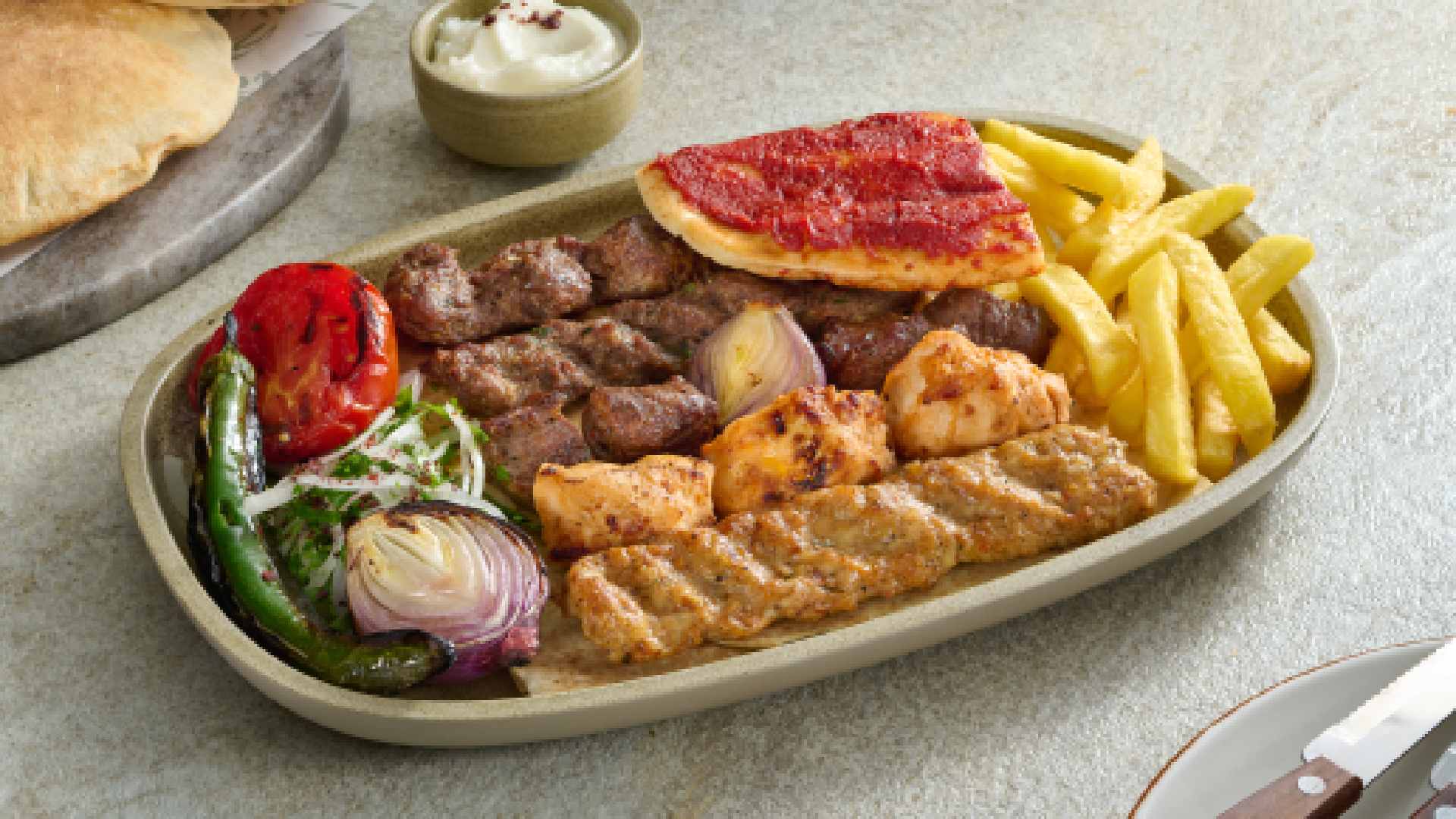 AL HALLAB mixed grill platter featuring grilled lamb cubes, chicken skewers, kafta, fries, grilled tomato, onions, flatbread, and garlic sauce on a serving plate.
