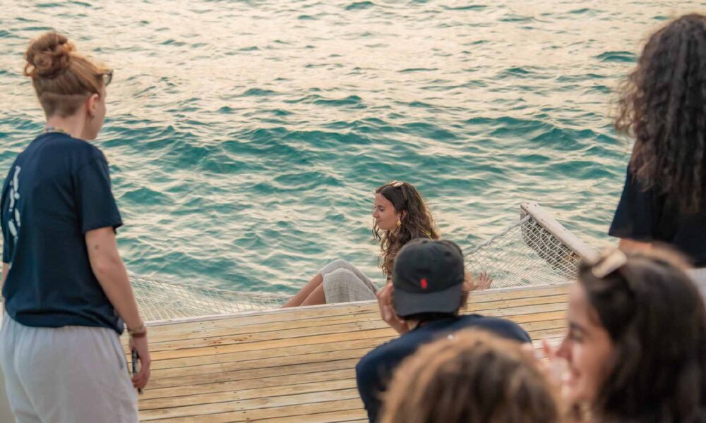 Sun Siyam team members and visiting students sit and stand on a wooden deck by the ocean, observing a marine conservation activity using a floating net in the lagoon.