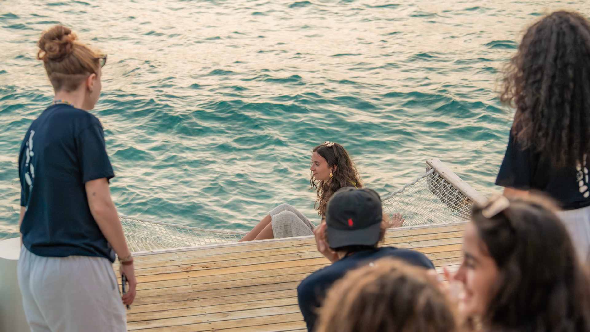 Sun Siyam team members and visiting students sit and stand on a wooden deck by the ocean, observing a marine conservation activity using a floating net in the lagoon.