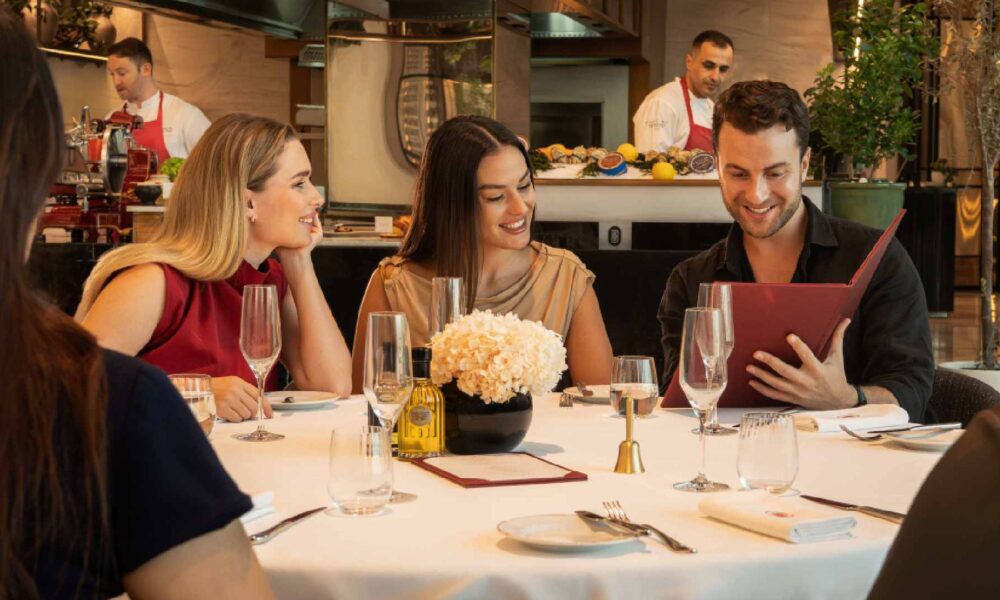 Guests seated around a white‑clothed table at Brasserie Boulud dine together while reviewing the menu, with champagne glasses, a floral centrepiece, and chefs working at the open kitchen counter in the background.