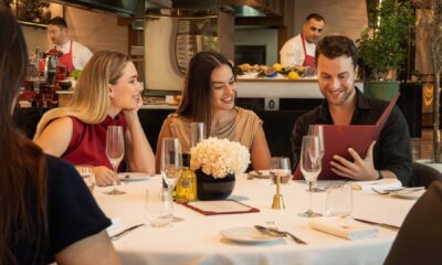 Guests seated around a white‑clothed table at Brasserie Boulud dine together while reviewing the menu, with champagne glasses, a floral centrepiece, and chefs working at the open kitchen counter in the background.