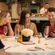 Guests seated around a white‑clothed table at Brasserie Boulud dine together while reviewing the menu, with champagne glasses, a floral centrepiece, and chefs working at the open kitchen counter in the background.