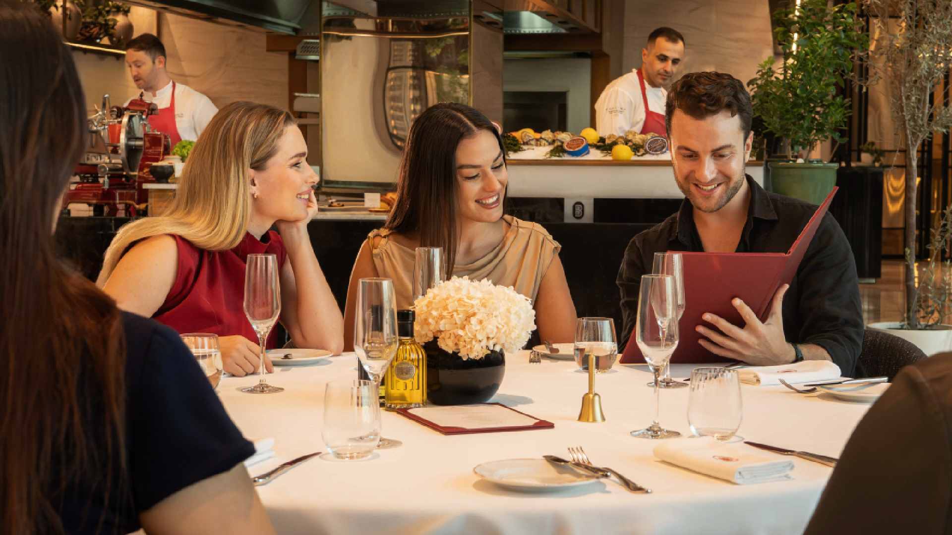 Guests seated around a white‑clothed table at Brasserie Boulud dine together while reviewing the menu, with champagne glasses, a floral centrepiece, and chefs working at the open kitchen counter in the background.