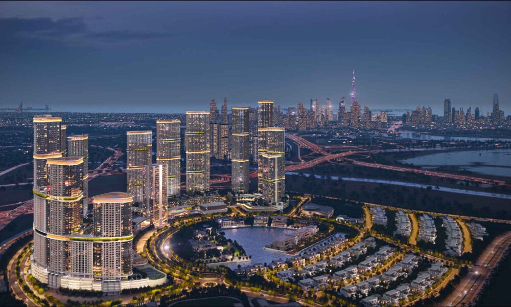 Aerial night view of Sobha Realty master-planned waterfront community featuring illuminated high-rise towers, residential villas, landscaped roads, and Dubai skyline in the background