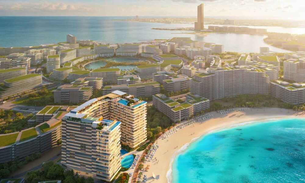 Aerial view of the ELEVATE coastal masterplan showing modern mid-rise buildings with green rooftops surrounding lagoons, beachfront residences with swimming pools, a sandy beach, and turquoise sea.