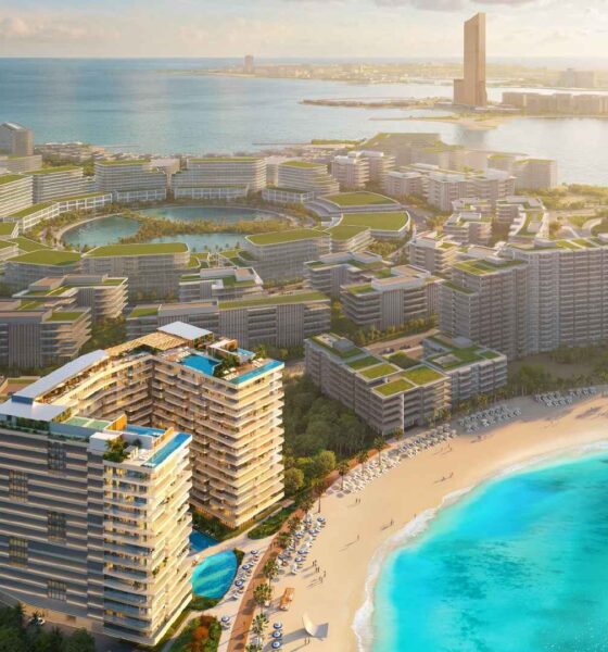 Aerial view of the ELEVATE coastal masterplan showing modern mid-rise buildings with green rooftops surrounding lagoons, beachfront residences with swimming pools, a sandy beach, and turquoise sea.